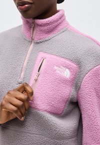 Persona che indossa una giacca in pile grigia e rosa con tasca sul petto con cerniera e collo alto, mano che aggiusta la cerniera.