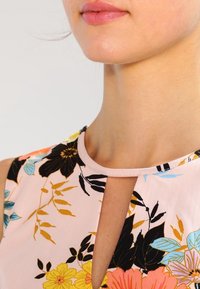 Lähikuva naisen alahuulesta ja kaulasta, jolla on päällä ilman hihoja kukallinen toppi avosylinterillä, pehmeässä vaaleanpunaisessa ja elävissä väreissä.