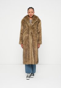 Μακριά ψεύτικη γούνα σε ανοιχτό καφέ χρώμα, με φαρδύ γιακά, μπροστινό κλείσιμο και παχύ υλικό. Συνδυασμένη με μπλε καμπάνα τζιν και μαύρα αθλητικά παπούτσια.