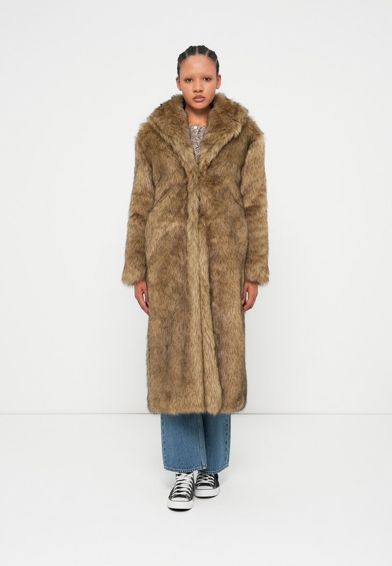 Μακριά ψεύτικη γούνα σε ανοιχτό καφέ χρώμα, με φαρδύ γιακά, μπροστινό κλείσιμο και παχύ υλικό. Συνδυασμένη με μπλε καμπάνα τζιν και μαύρα αθλητικά παπούτσια.