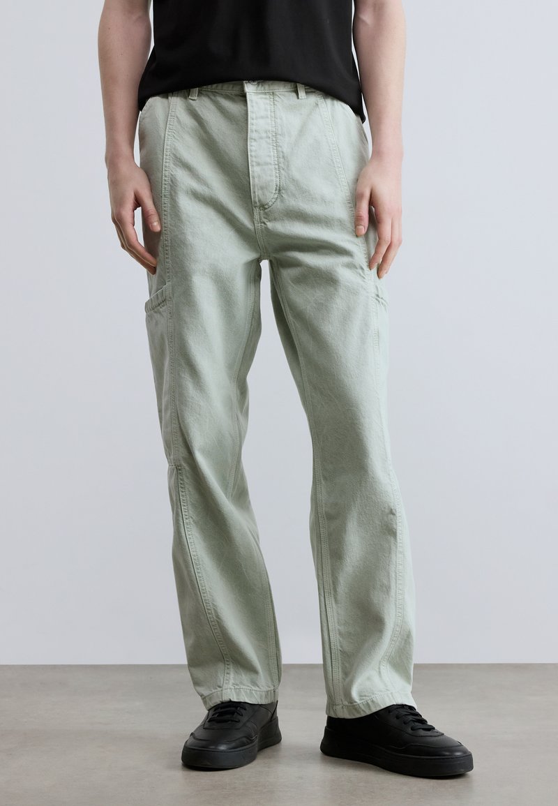 Φόρμες cargo ανοιχτού πράσινου με χαλαρή εφαρμογή, με εμπρόσθιες τσέπες και υφασμάτινη υφή. Συνδυάζονται με μαύρα αθλητικά παπούτσια.