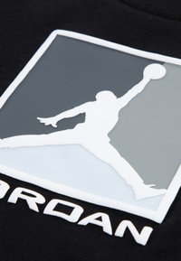 Μαύρο ύφασμα με ανάγλυφο λευκό λογότυπο που απεικονίζει μια φιγούρα σε άλμα και τη λέξη «JORDAN» σε λευκό από κάτω, τοποθετημένα πάνω σε γκρι φόντο.