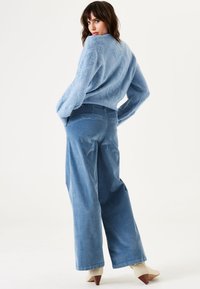 Helles blaues Strickpullover mit strukturiertem Muster und weiten Ärmeln, kombiniert mit locker sitzenden hellblauen Cordhosen und beigen, hohen Stiefeletten.
