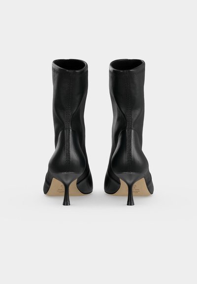 Paire de bottines en cuir noir à talons aiguilles fins, vues de dos sur un fond clair uni.