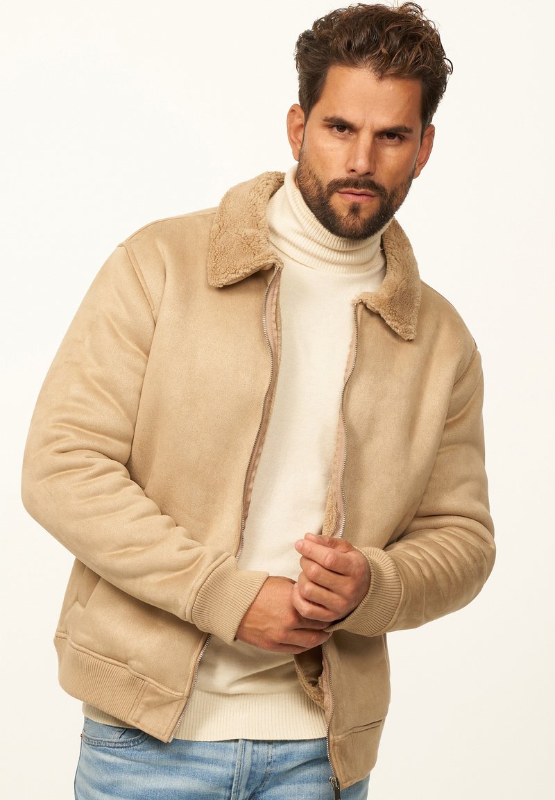 Beige Bomberjacke mit Schafwollfutter, Reißverschluss, gerippten Bündchen und Kragen, mit glatter, suede-ähnlicher Textur, getragen über einem hellen Rollkragenpullover.