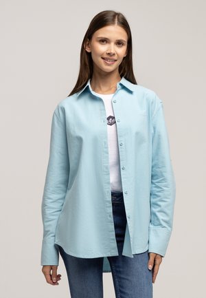 Jasnoniebieska koszula z długim rękawem, wykonana z miękkiego materiału, z zapięciem na guziki z przodu, w luźnym fasonie i kołnierzykiem typu spread. Nosi się ją na białej koszulce.