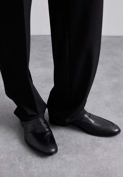 Schwarze Lederloafers mit glatter Textur, ausgestattet mit einem eleganten Design und auffälligen Riemen-Details, kombiniert mit maßgeschneiderten schwarzen Hosen.