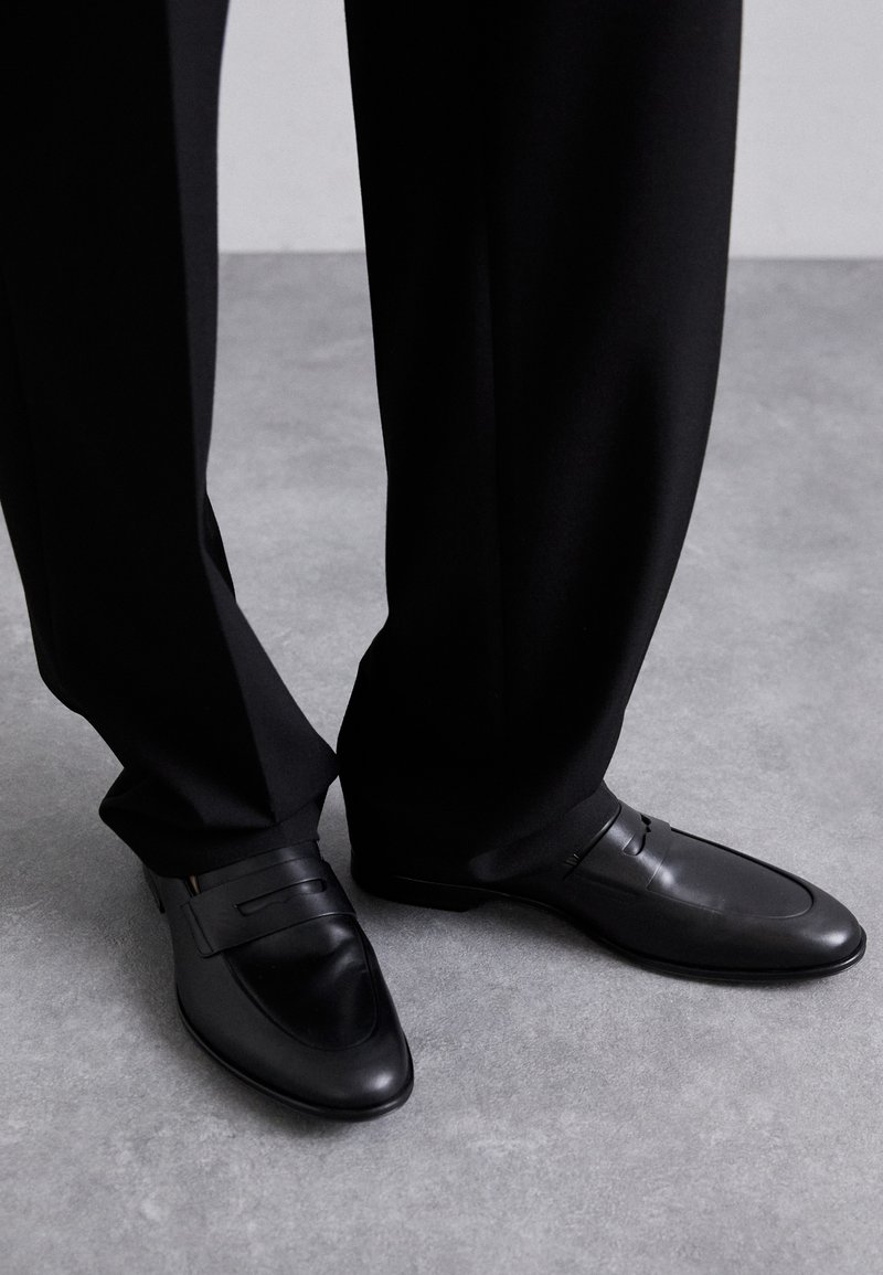 Black leather loafers with a smooth texture, featuring a sleek design and prominent strap detail, paired with tailored black trousers.