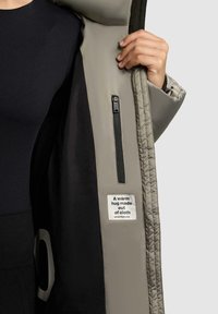 Gepolsterte graue Jacke mit schwarzem Innenfutter, ausgestattet mit einer Reißverschlusstasche und einem Etikett mit der Aufschrift "Eine warme Umarmung aus Stoff."