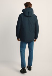 Granatowa pikowana kurtka z kapturem, o gładkiej fakturze i luźnym kroju; w zestawieniu z niebieskimi spodniami dżinsowymi i czarnymi butami.