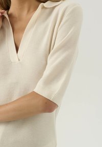 Žena v krémovém krátkém rukávu pleteném polo tričku s hlubokým výstřihem do V, zobrazená od krku po střed paže.