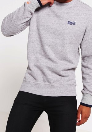 Homme portant un sweat-shirt gris clair à col rond avec un logo bleu marine sur la poitrine et un pantalon noir, posant avec une main près du menton.