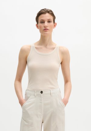 Femme aux cheveux bruns courts portant un débardeur côtelé beige et un pantalon taille haute de couleur claire, debout avec les mains dans les poches devant un fond blanc.