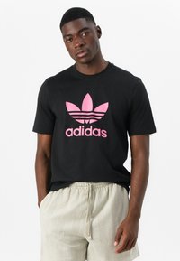 Svart bomull T-skjorte med en stor rosa Adidas-logo med tre horisontale striper. Korte ermer og klassisk rund hals-design.