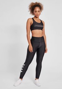 Czarny sportowy krótki top z logo „HUMMEL” oraz legginsy z wysokim stanem, ozdobione białymi wzorami w kształcie szewronu po bokach. Teksturowany, dopasowany fason.