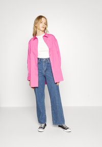 Camisa rosa oversized, t-shirt branca, jeans azuis de cintura alta, ténis pretos. Textura suave, corte descontraído, roupa casual.