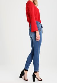 Blusa roja de manga larga con puños acampanados, jeans ajustados azules con cinturón y sandalias negras de tacón alto con tiras en el tobillo.