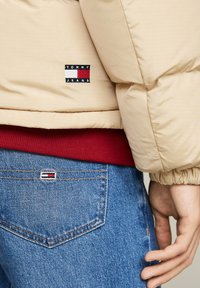 Beige puffer jacket with a red ribbed hem, featuring a black, red, and white "TOMMY JEANS" logo. Blue denim jeans with a similar logo patch.