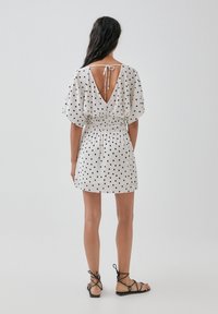 Puku puntini valkoisena ja mustana, jossa on syvä V-mallinen selkä, joustava vyötärö ja löysät hihat, yhdistetty mustiin remmisandaaleihin.