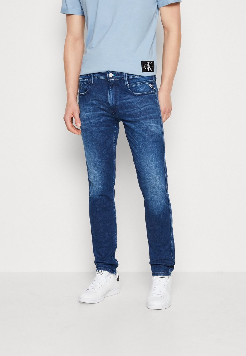 Homme portant un jean slim bleu, un t-shirt bleu clair avec une étiquette noire et des baskets blanches, debout devant un fond clair uni.