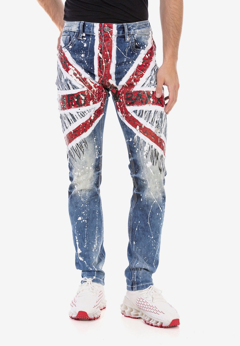 Jeans en denim bleu caractéristiques d'un design de Union Jack en rouge et blanc, avec des détails de éclaboussures de peinture et un texte brodé sur le devant.