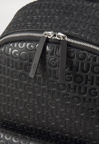 Mochila negra con un material texturizado que presenta el logotipo "HUGO" en relieve, cremallera metálica con tiradores y un diseño liso.