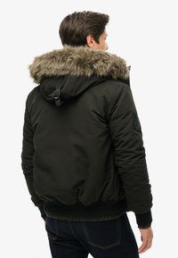 Veste matelassée vert foncé avec une capuche en fausse fourrure, des poignets élastiques et un ourlet côtelé. Comprend un détail de patte sur le côté de la manche.
