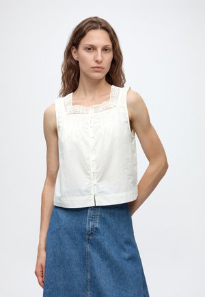 Ung kvinde iført en hvid ærmeløs bluse med blonde detaljer og et blåt cowboyskørt, stående mod en ensartet lys baggrund.