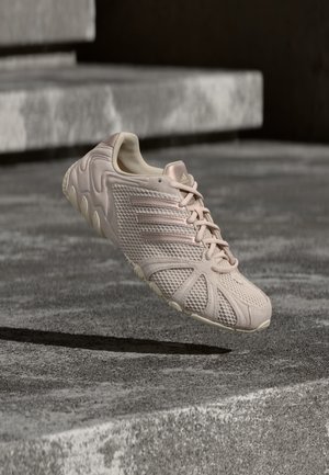 Jasnoróżowy sneaker Adidas z siateczkową cholewką, trzema paskami z boku, zawiązanymi sznurowadłami, zawieszony nad chropowatymi betonowymi schodami.