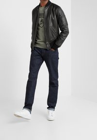 Chaqueta bomber de cuero negro, sudadera verde con logo, vaqueros oscuros y zapatillas blancas. La chaqueta tiene un bolsillo con cremallera y puños acanalados.