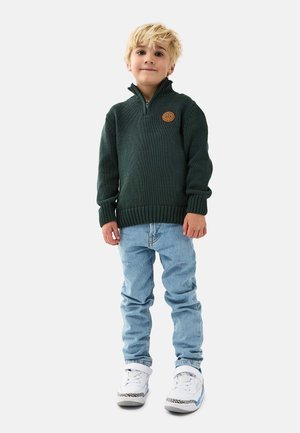 Grüner gestrickter Pullover mit hohem Kragen und gerippten Bündchen, kombiniert mit hellblauen Jeans und weißen Sneakers mit schwarzen Akzenten.