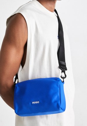 Personne portant une chemise sans manches blanche et un sac bandoulière Hugo bleu vif avec une sangle noire réglable sur l'épaule.