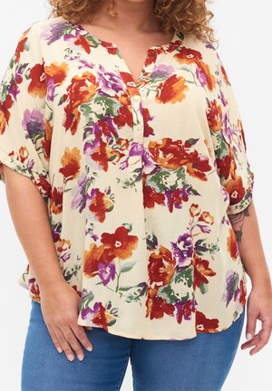 Frau trägt eine cremefarbene Bluse mit rotem, lila und orangefarbenem Blumenmuster, kurzen Ärmeln und blauen Jeans, zeigt ein teilweises Unterarmtattoo.