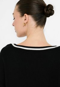 Zwarte trui met een brede wit gestreepte halslijn, zichtbaar een gladde brei-textuur. Gouden stud oorbellen zijn zichtbaar tegen een lichte achtergrond.