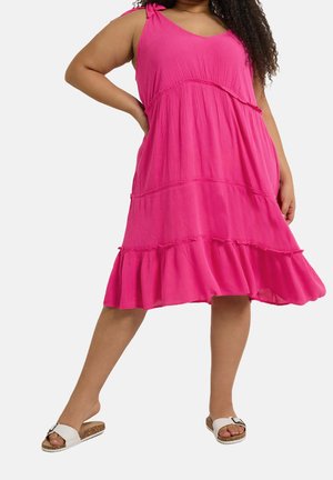 Frau trägt ein ärmelloses, knielanges Kleid in leuchtendem Pink mit Volant-Saum und weiße Sandalen ohne Absatz, steht mit der Hand auf der Hüfte.