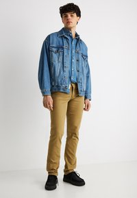 Νέος άνδρας που φοράει μπλε τζιν μπουφάν, μπλε τζιν πουκάμισο, παντελόνια μουσταρδί και μαύρα αθλητικά παπούτσια, στέκεται μπροστά σε λευκό φόντο.