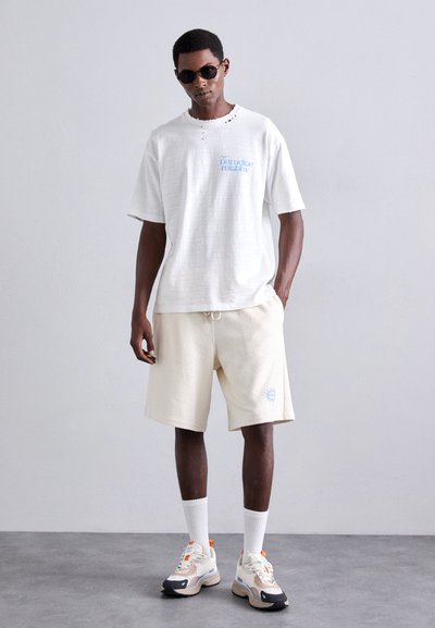 Hvit teksturert t-skjorte med blå grafikk, beige teksturerte shorts med brodert sol-logo. Klumpete joggesko med fargerike detaljer.