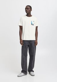Camiseta de algodón en blanco roto con un bolsillo estampado en azul y verde, combinada con pantalones vaqueros oscuros y holgados de color gris y zapatillas grises.