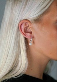 Silverfärgade hoops med en liten, hängande pärla. Hoopen har en texturerad, glittrande acccent. Håret är ljust och något vågigt.