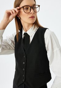 Svart skräddarsydd väst med sex knappar och en ficka, bärs över en vit skjorta med krage och svart slips. Modellen har långt hår och glasögon.