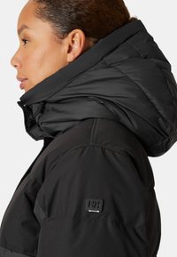 Zwarte pufferjack met hoge kraag, voorzien van een zachte textuur en een logo-label op de linker mouw. Het ontwerp benadrukt warmte en comfort.