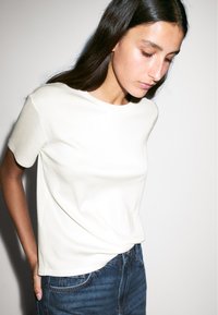 T-shirt di cotone di colore avorio, maniche corte, colletto rotondo, texture morbida, abbinata a jeans in denim blu scuro, design minimalista, senza fantasie.