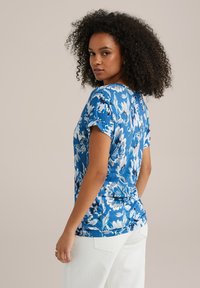 Zila ziedu T-kreklā ar īsām piedurknēm, ar noapaļotu kakla izgriezumu un vieglu, teksturētu audumu. Dizainā ir iekļauti balti un tumši zili ziedi.