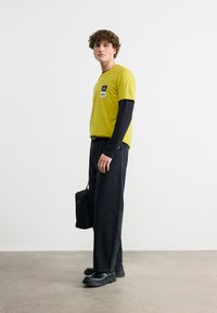 Jeune homme portant une chemise jaune à manches courtes par-dessus un haut noir à manches longues, pantalon noir large, chaussures noires, tenant un sac noir, debout contre un mur blanc.