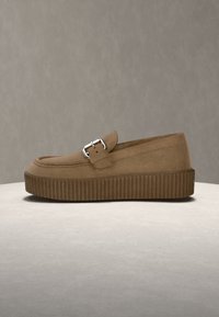 Mocassin compensé en daim marron avec boucle argentée et semelle en caoutchouc rainurée sur une surface unie.