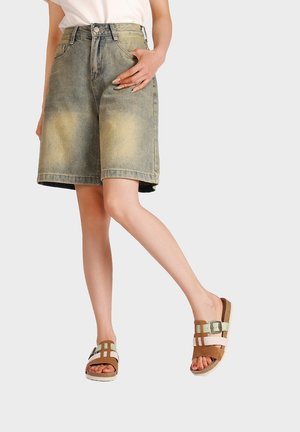 Persona che indossa pantaloncini di denim sbiaditi fino al ginocchio e sandali marroni slip-on con fibbie, in piedi con una gamba accavallata.