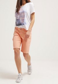 Shorts de mezclilla naranja ligeros y cortos con un botón frontal y bolsillos, combinados con una camiseta gráfica blanca y zapatillas grises sin cordones.