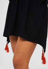 Black dress with a textured fabric, featuring an asymmetrical hem and orange tassels with metallic accents at the lower sides.