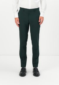 Sötétzöld, sima anyagból készült szabott nadrág, slim fit fazonnal és lapos elülső résszel, fekete cipővel és szürke zoknival párosítva.