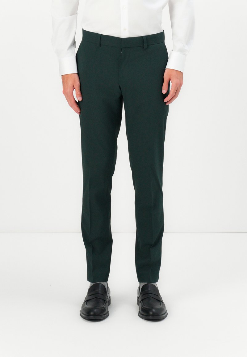 Sötétzöld, sima anyagból készült szabott nadrág, slim fit fazonnal és lapos elülső résszel, fekete cipővel és szürke zoknival párosítva.